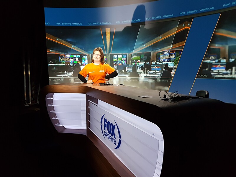 Lize sitting behind the FOX sports news desk with her hands on her hips and smiling.