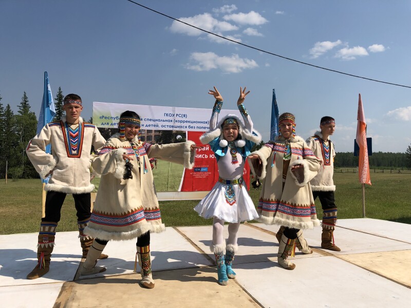 Five young people in traditional costumes pose on a stage with grass and a blue sky in the background.