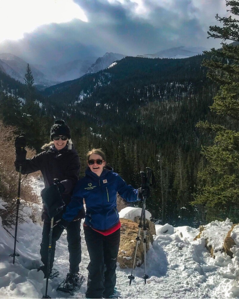 Two women stand in the mountains. They're wearing snowshoe equipment and smiling for the camera.