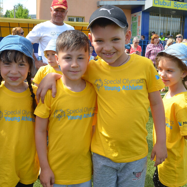 Three smiling young children in yellow Special Olympics Young Athletes t-shirts face the camera. They are standing on a grass pitch with a crowd behind them. 
