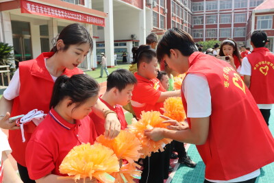 Volunteers in red vests assist participants during an outdoor Special Olympics activity, helping distribute and organize yellow pom-poms on a school playground. Several participants and volunteers work together in front of a school building during a community engagement event.