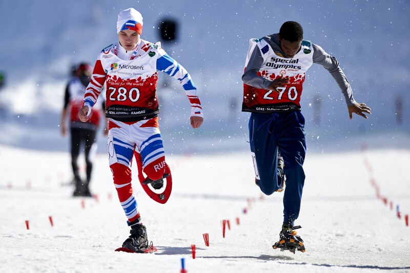 Two male athletes running on a snowy surface towards the camera in snowshoes.