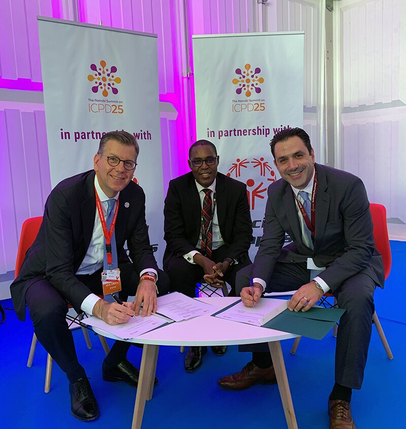 Three men sitting at a table; each man on either side of the middle man are signing papers. Signage in the background reads: ICPD25 in partnership with Special Olympics.