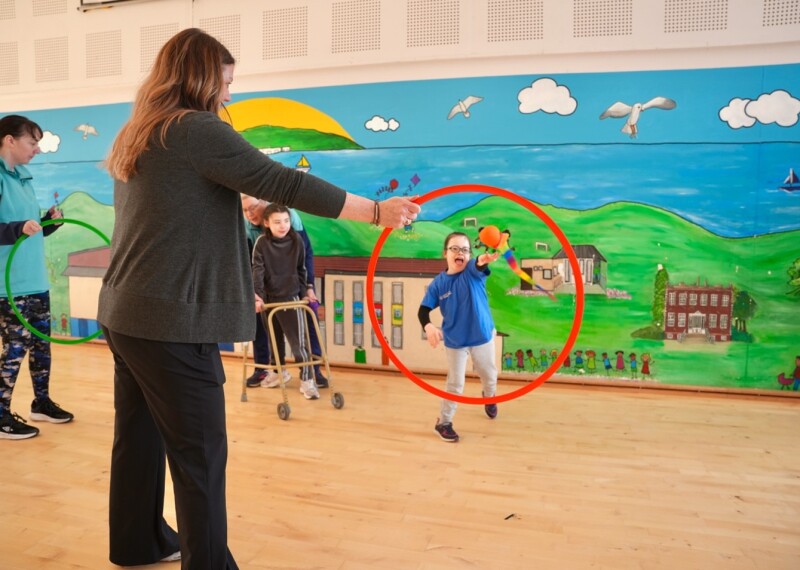 In the foreground, a woman stands holding a red hoop while a child throws a orange ball with a multicolor tail through it. Another child using a mobility walker is nearby. The room has a wooden floor and a brightly painted mural on the wall depicting hills, buildings, clouds, birds, and water.