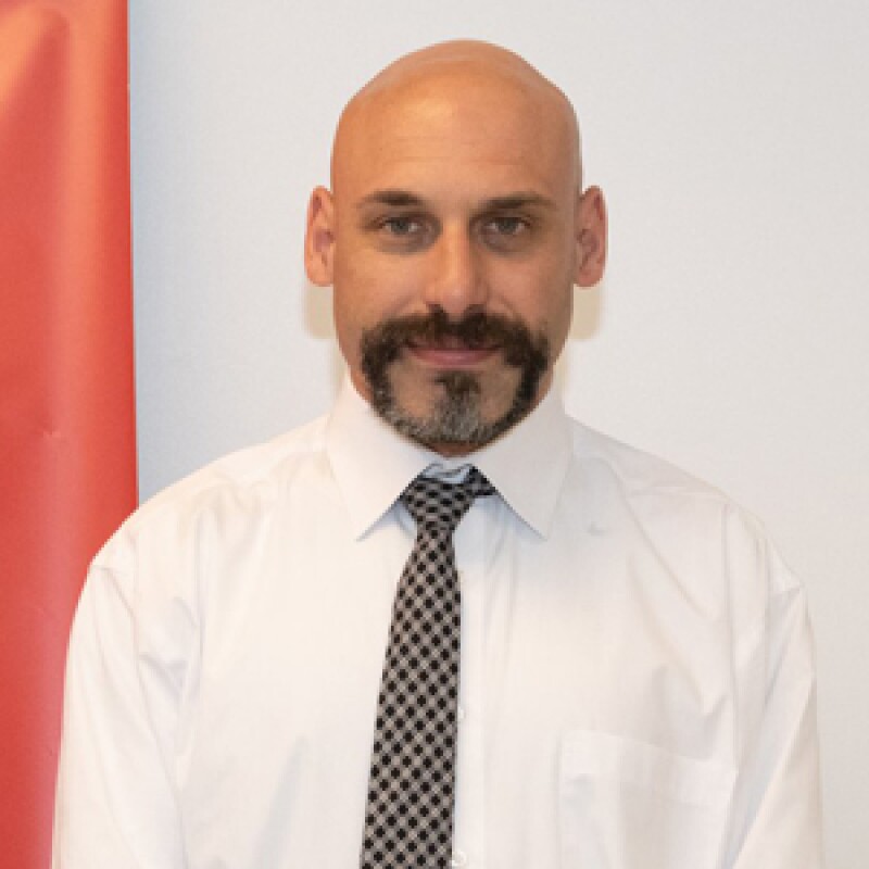 Paul Asaro smiling for a portrait photo. He's wearing a white button up dress shirt and a black and white diamond checkerboard tie.