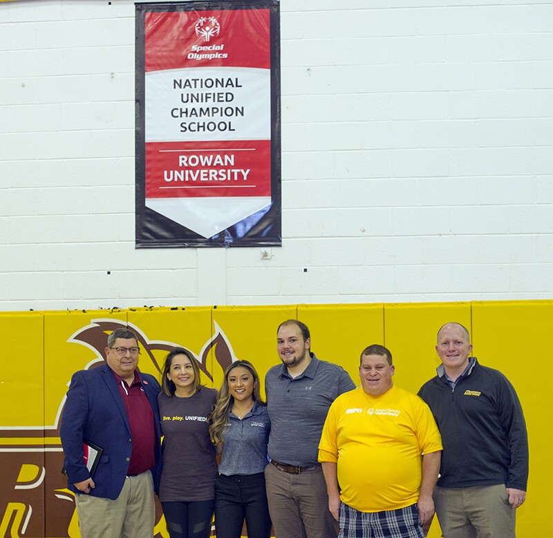 A group of 6 young adults and Rowan and EPSN representatives stand under a Rowan University Unified Champion Schools banner in a gymnasium.
