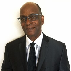 Robert "Bob" Beamon in a black suite and tie with a white and black vertical striped collared shirt.
