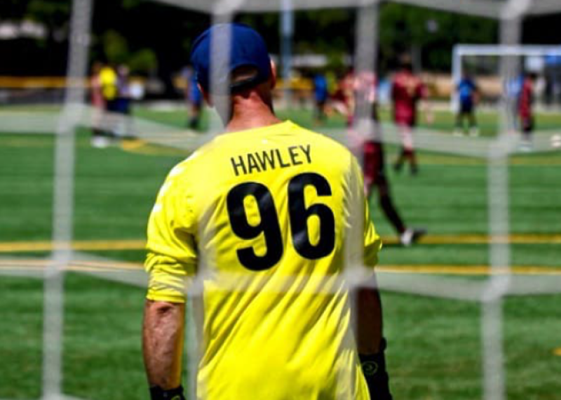 A Special Olympics athlete wearing a yellow goalie jersey stands in the soccer net, facing the field. 