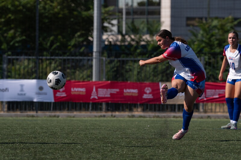 A woman executes a powerful kick, sending a soccer ball flying across a field, surrounded by green grass.