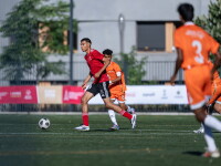 A football player in a red jersey dribbles the ball upfield as opponents in orange jerseys pursue him.