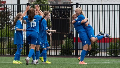 Group of girls wearing a blue uniform celebrating on a football field.
