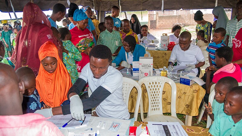 a man with examination gloves on is performing a test while athletes and other individuals watch under an outdoor tent.
