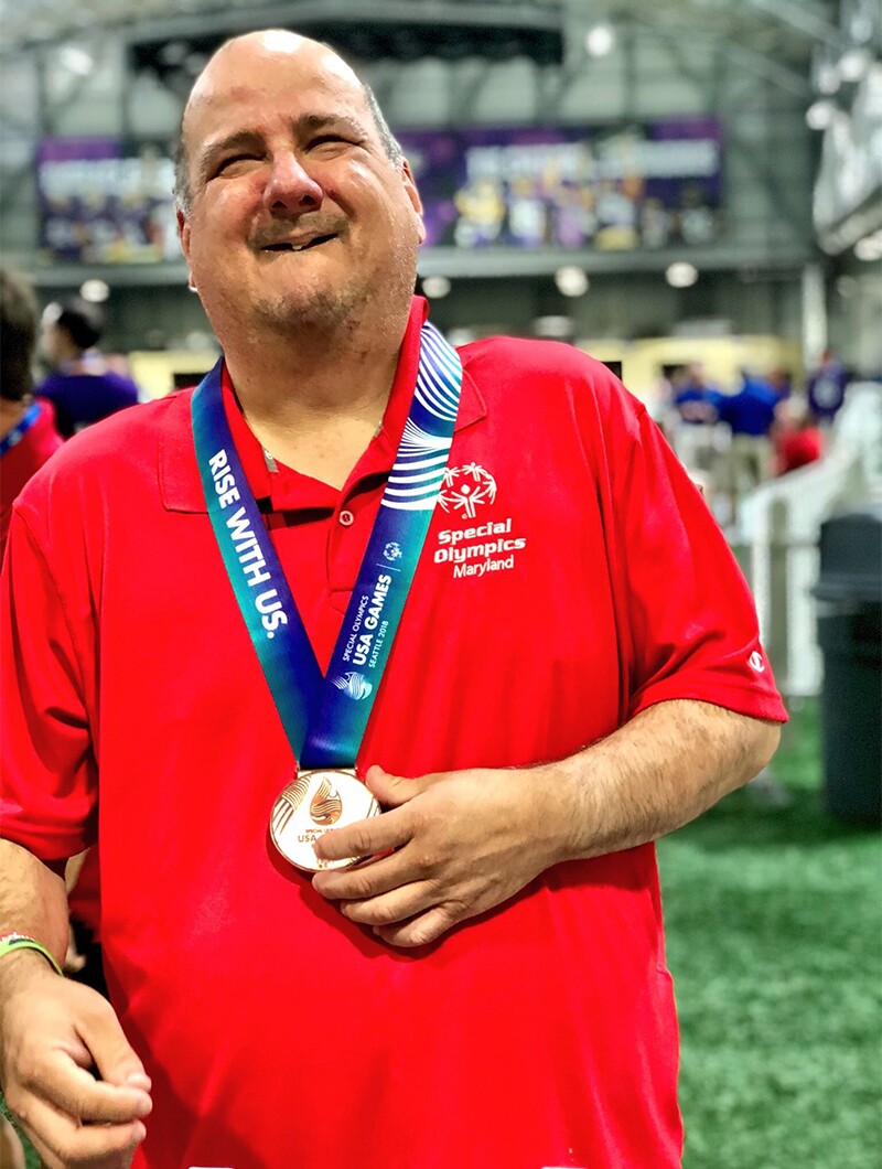 Ben after winning Bronze at the 2018 Special Olympics USA Games in Seattle. He is in standing inside the facility with patrons and athletes in the background; Ben is wearing a red Special Olympics polo and has a Bronze medal around his neck.