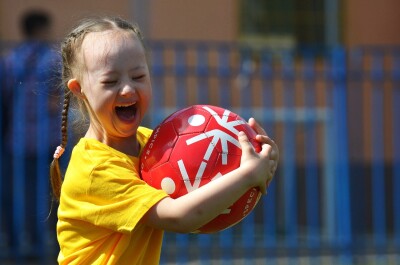 A young girls in a yellow t-shirt holds a red football. Her eyes are closed and she is shouting in glee.