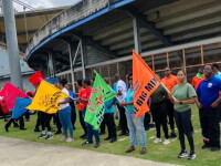 A group of people stands outdoors along a walkway beside a stadium or large sports venue. Several participants hold brightly colored flags, including orange, green, yellow, blue, and red flags. The orange flag in the foreground displays the text “THE BIG MIX.”