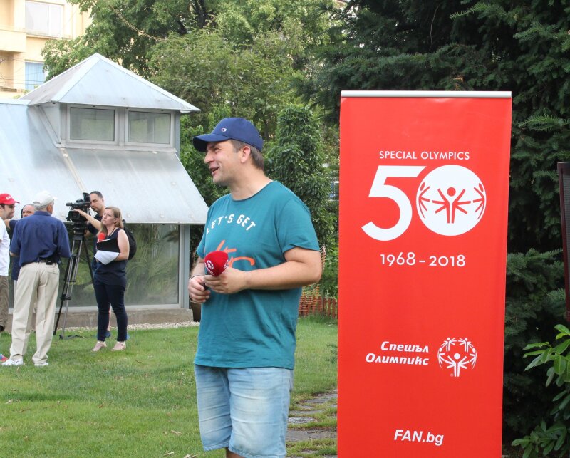 A man wearing a baseball hat, a turquoise t-shirt and jeans shorts stands in front of a red Special Olympics 50 pull-up banner holding a red microphone in a garden setting.