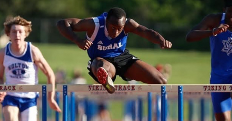 A male athlete leaps over a hurdle on the track. He is wearing a blue track uniform.