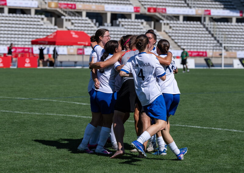 A group of women soccer players wearing white and blue uniforms celebrate in a team huddle on the field of a large stadium with mostly empty stands in the background. 