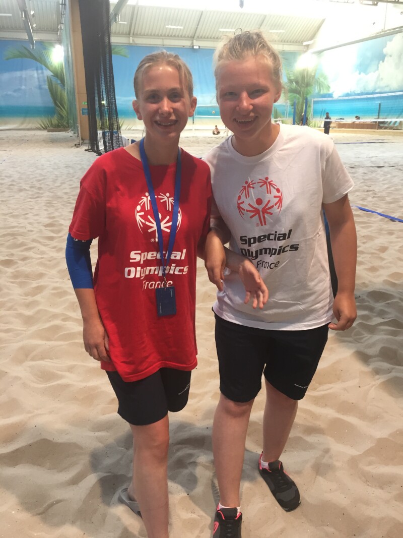 Two blonde girls in Special Olympics t-shirts stand side by side smiling at the camera standing on sand with a beach volleyball net in the background.