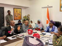 A group of about ten people seated around a conference table during an official meeting in a boardroom. Several participants are speaking and listening, with microphones, notebooks, and documents placed on the table. Two red balls with the Special Olympics logo sit at the center. A national flag of Niger stands upright behind the group, and framed artwork is visible on the walls.