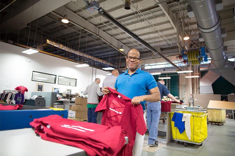 Bank of America Support Services print shop produces all in house t-shirts. Employee in a blue polo holding a red t-shirt from a stack of red t-shirts in front of him.