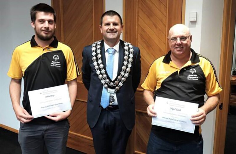 Three men, two holding certificates and the man in the middle with an award around his neck. 