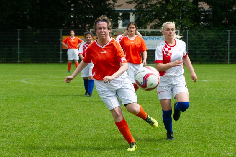 Women in football/soccer jerseys run after a football on a green pitch.