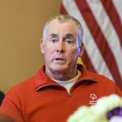 John C. McGinley in a red zip up Special Olympics sweater with the American flag in the background.