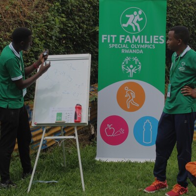 One man drawing on a white board and another standing next to a pull up banner that reads, Fit Families Special Olympics Rwanda.
