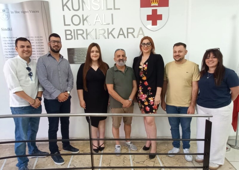 Seven people in casual dress, four men and three women, pose for a photograph in front of a wall with the Birkirkara city crest