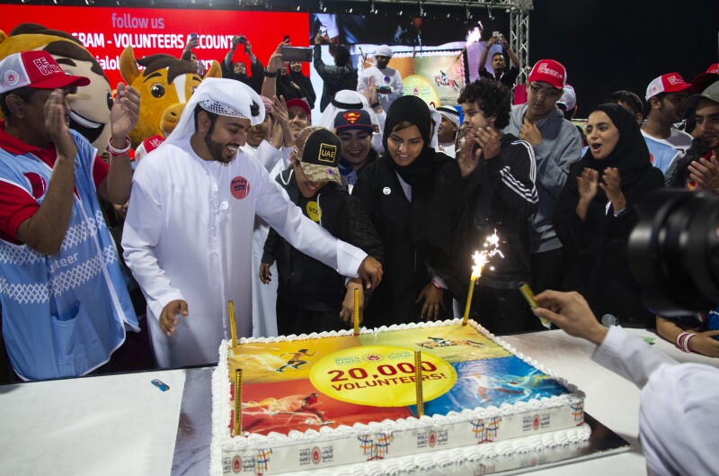 A group of people stand around smiling and clapping as a man lights the candles on a large rectangular cake with the words '20,000 volunteers' on it.