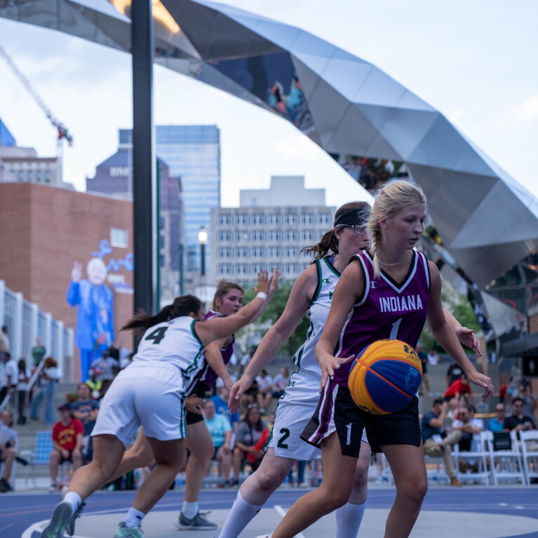 Young women on the basketball court, one person dribbles and is followed by a player on the opposing team. 