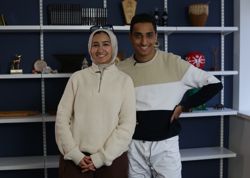 Young man and woman standing in front of a bookshelf with nicknacks on it. 