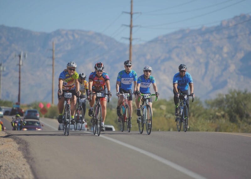 A group of 6 cyclists riding down a road in a rural area.