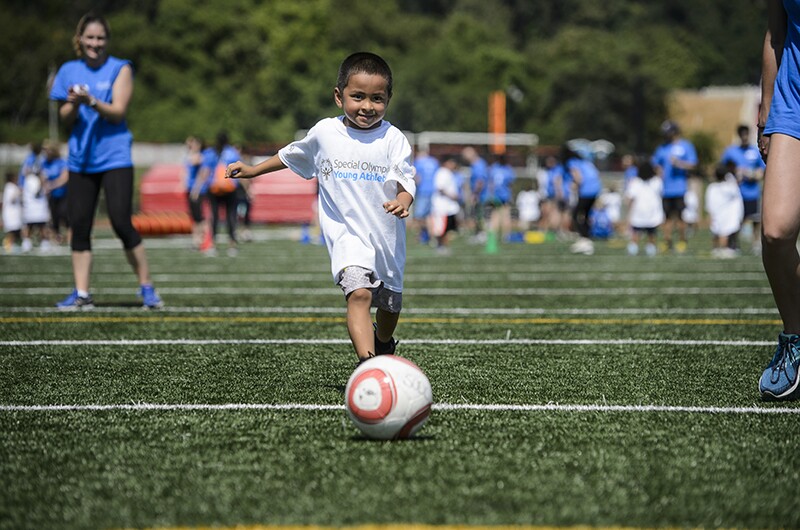 A young athlete in the center smiles at the camera as he prepares to kick a soccer ball on a soccer field while two volunteers watch.