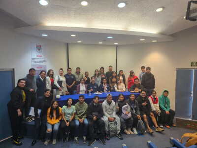 Large group of approximately 30 people gathered in a classroom or meeting room, posing for a group photo around a long table covered with a blue tablecloth. Some individuals are seated in the front row while others stand behind them. The room has white walls, recessed ceiling lights, and two doors on either side. A banner on the wall includes the words “Misión” and “Visión” in Spanish.