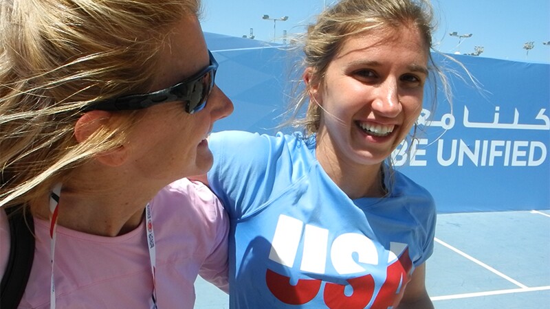 Brittany with a woman on the courts. Brittany is smiling big and has on a USA shirt. in the background is a Play Unified sign.