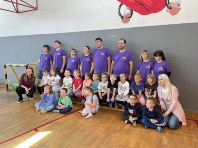 A group picture with adults lined in purple shirts standing and younger students/athletes sitting down. The picture is taken in a basketball court with a grey wall backdrop behind them
