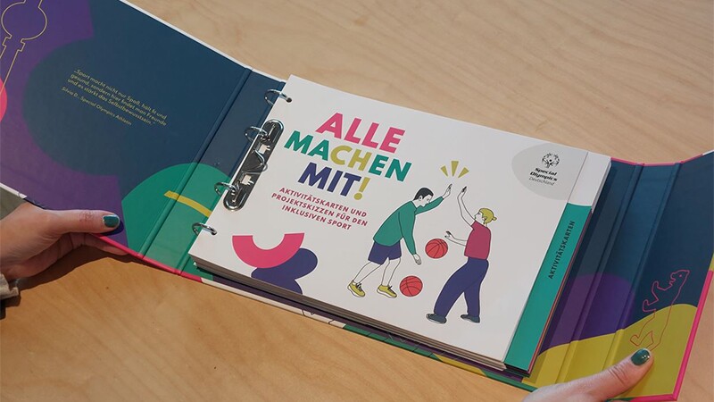 Shows two hands holding Alle Machen Mit ring binder.  It is opened on the first page of the folder contents, showing Alle Maching Mit! text and a drawing of two people bouncing basketballs while giving each other a high five.