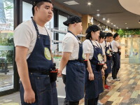 A line of trainees in matching white shirts, dark aprons, and black caps stand side by side inside a brightly lit restaurant or café. They are positioned along a wooden floor near a service counter, with tools or cleaning supplies attached to their waists. Large windows line the left side, and the interior features warm lighting, hanging fixtures, and decorative greenery in the background.