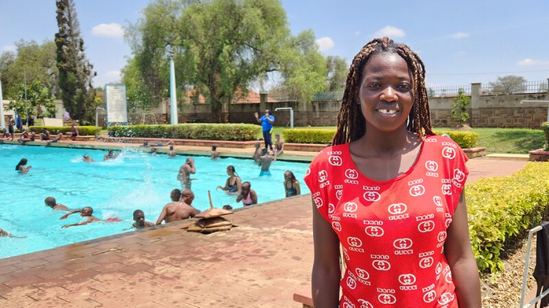 Coach Rehab Njunguna smiles at the camera while standing in front of a swimming pool filled with athletes.