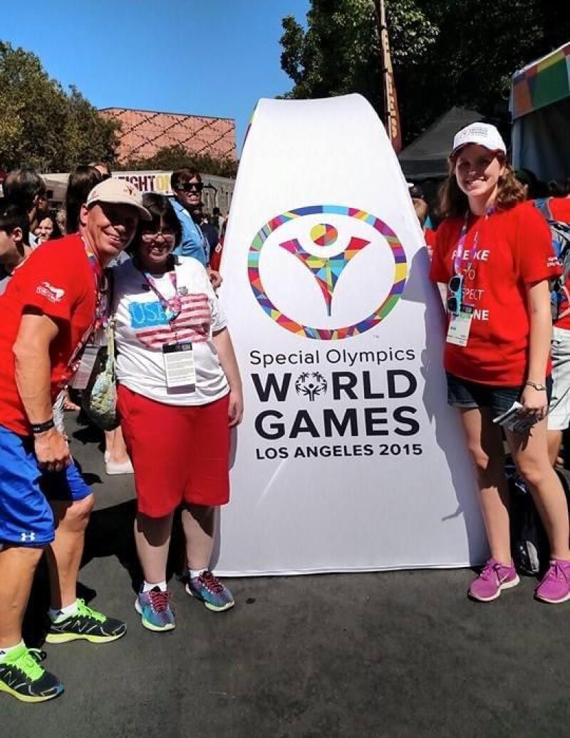 Three people pose in front of a large white sign that has the Special Olympics World Games LA 2015 logo on it. They are smiling.