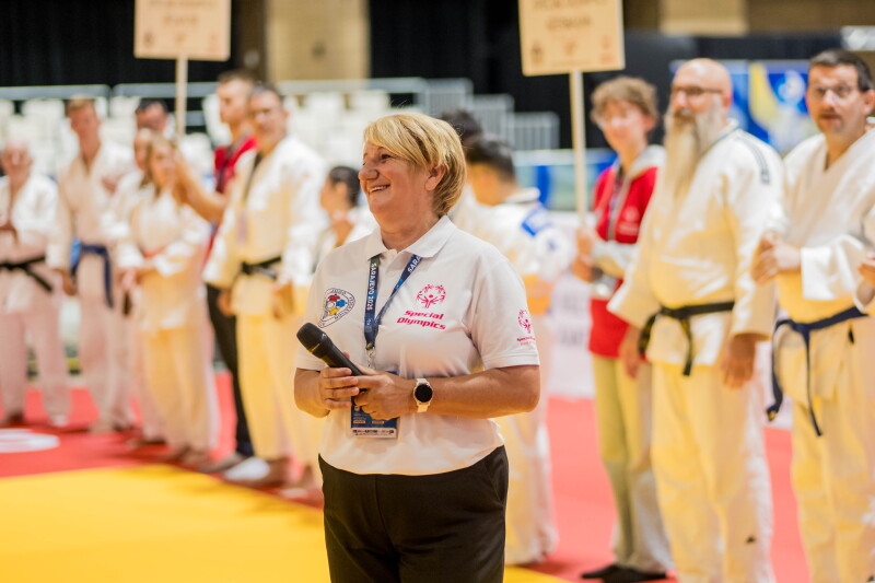 A woman standing on the tatatami, holding a microphone. Surrounded by athletes wearing the official judogi. 