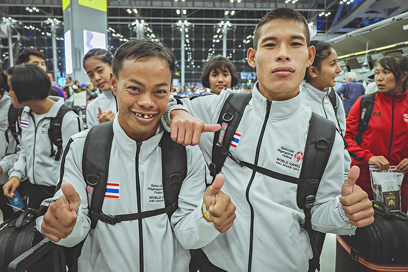 Nakorn Thiptabmak with another athlete giving the thumbs up. Both have on Special Olympics Thailand World Game Abu Dhabi 2019 zip up jackets. Athletes from Thailand re in the background.