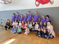 A group picture with adults lined in purple shirts standing and younger students/athletes sitting down. The picture is taken in a basketball court with a grey wall backdrop behind them