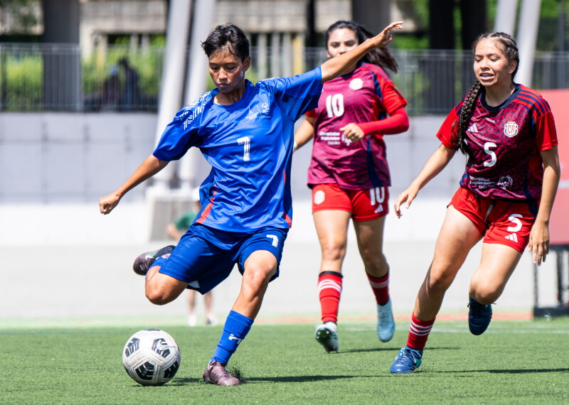 A male player prepares to kick the ball while two women from the opposing team come up behind him. 