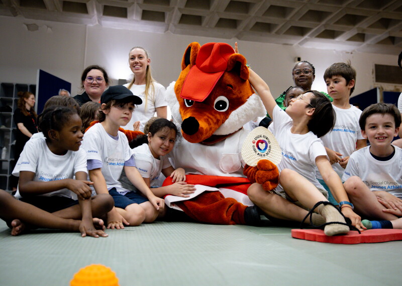 Children sit around a large fox mascot with a row of adults behind them.