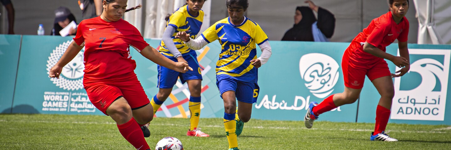 Four young women on the field playing football, two from each team with spectators in the background.