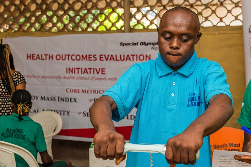 Young man holding an apparatus measuring his strength. A sign in the background reads: Health Outcomes Evaluation Initiative.