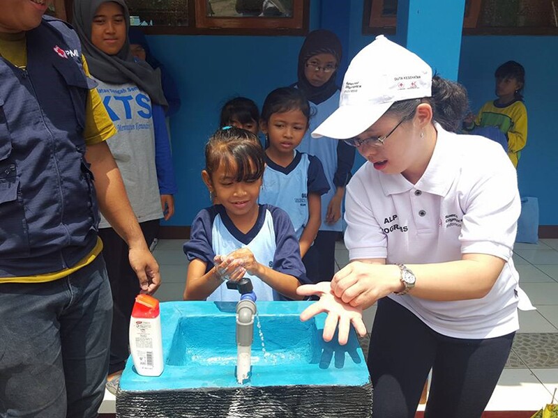 Young woman shows a young girl how to properly wash her hands while other young girls look on.